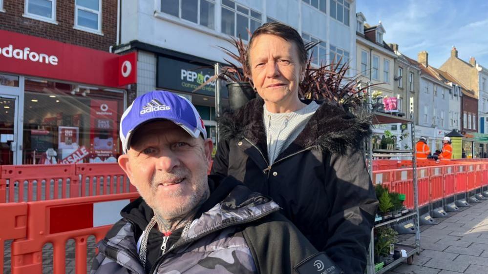 Jane Cole, on the right, with dark hair, wearing a light top and black coat, with her partner David Durrant, in a black and grey jacket and blue baseball cap. He is bearded and is using a wheelchair. They are pictured near some shops and a market flower stand, and some orange roadworks barriers