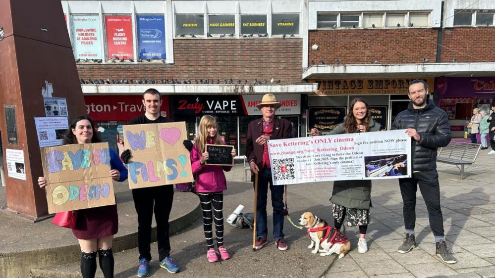 Six people, and a dog, stand in a town centre holding cardboard signs protesting against the closure of the Odeon cinema.