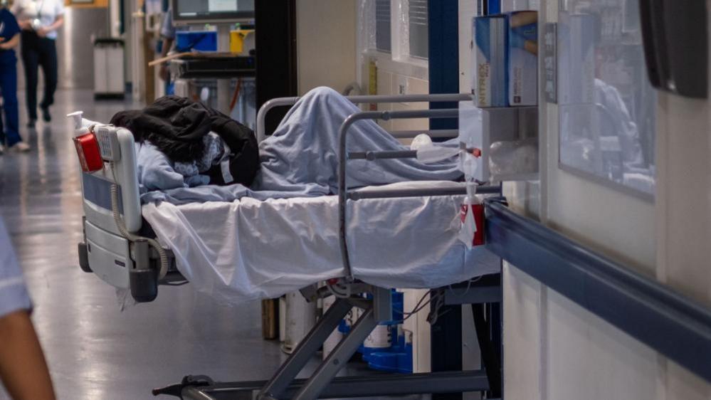 A picture of an NHS corridor. A patient hidden under a coat is lying on a hospital bed in a corridor. 