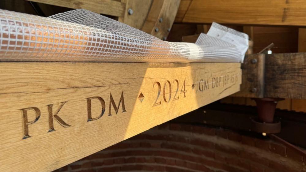 An oak beam, inside the cap of the Toft Monks Wind Pump, carved with the initials, PK, DM - the date year 2024 - GM, DBP, JBP, AS and JS. A sheet of reinforced plastic is over the top of the beam to protect it from the weather.