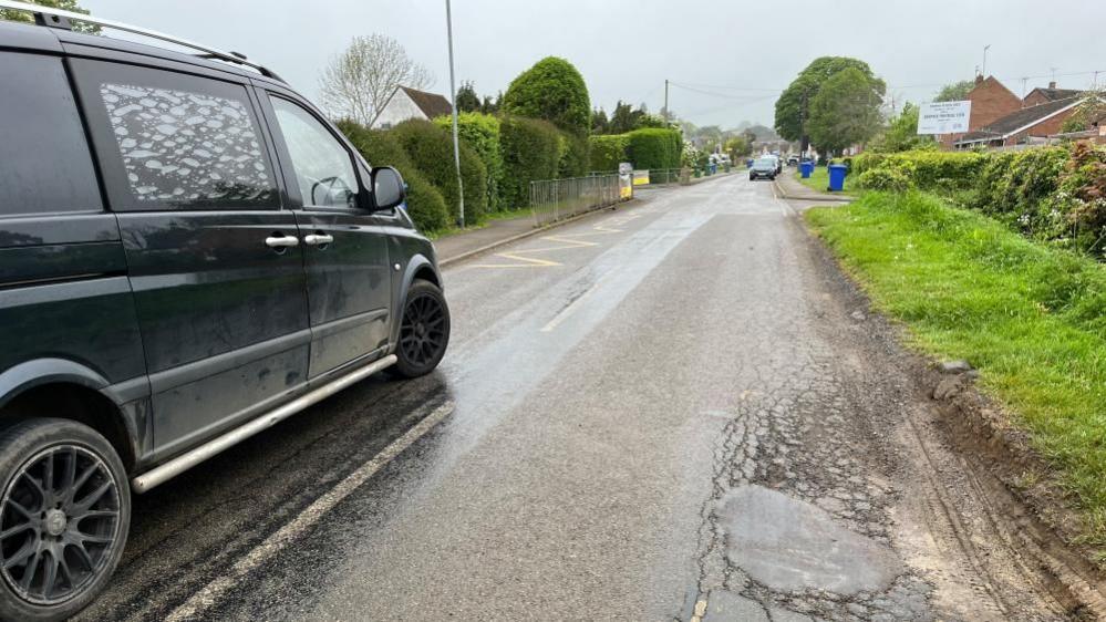 An MPV driving along a road into village with potholes showing