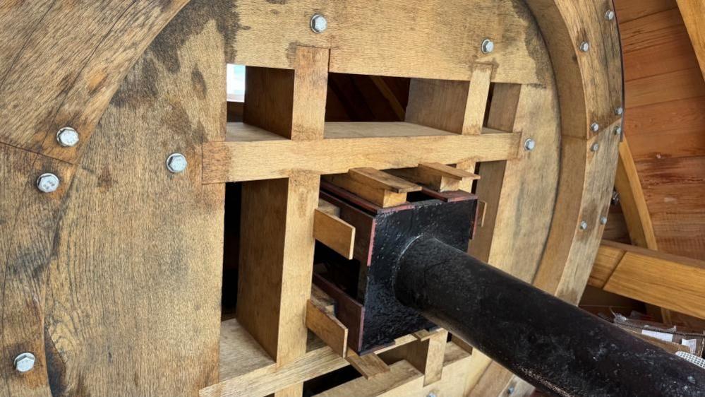 A wheel of oak, jointed and bolted together, and held onto a cast iron drive shaft with wooden wedges. The shaft is painted black, and part of the cedar clad cap is visible to the right of the image.