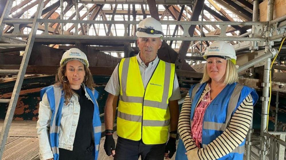 Two women are either side of one man, with all three wearing hard hats. The man is wearing a fluorescent jacket and the women are wearing blue and grey tops. 