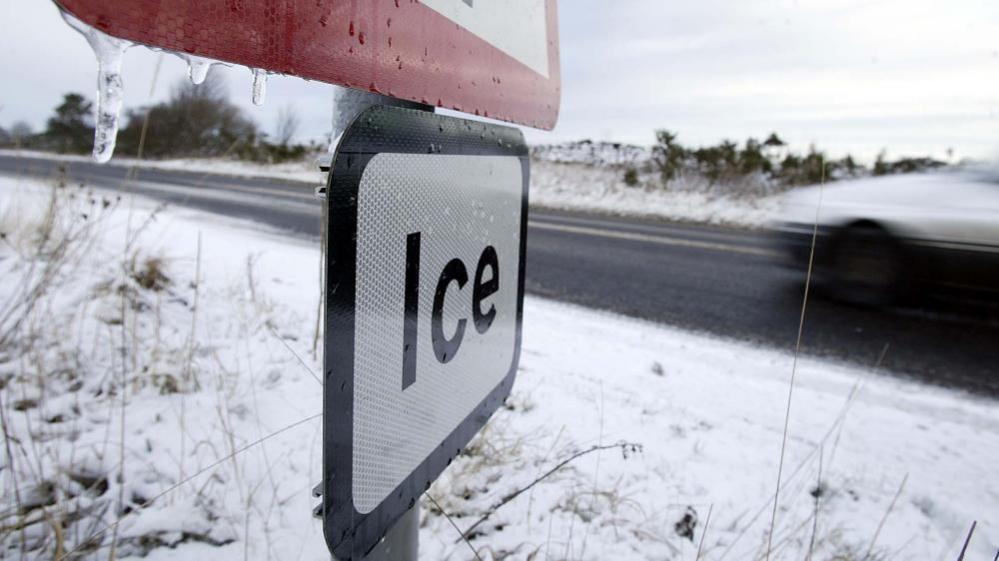 A road sign which says 'ice' can be seen next to a road with snow on either side of it. 