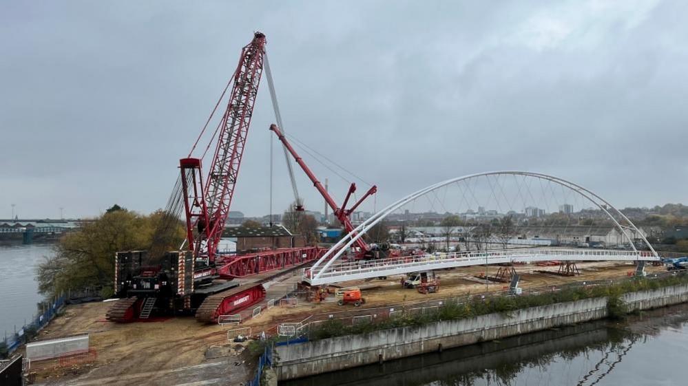 A view of the preparation work ahead of the bridge being lifted into place, with a large red crane on the left and a pre-assembled white bridge structure on the right.