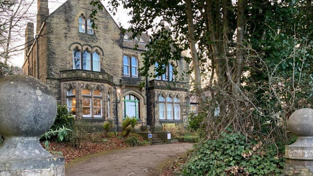 A large old brick building stands at the end of a driveway. The front garden is full of trees and lights can be seen through the large front windows. 