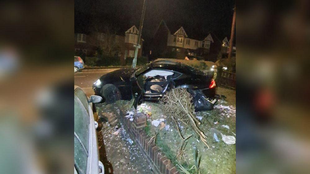 A black car with its side door open is partly in a front garden, where a tree is seen damaged alongside it and a part of a low wall has been knocked down. It is dark and there are semi detached houses seen in the background