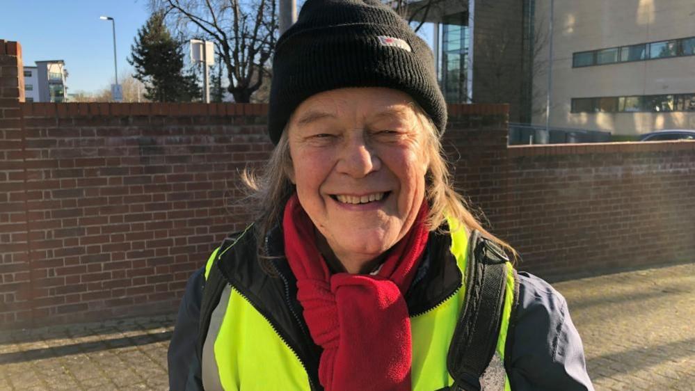 A man is smiling. her is wearing a hat and high visibility jacket. 