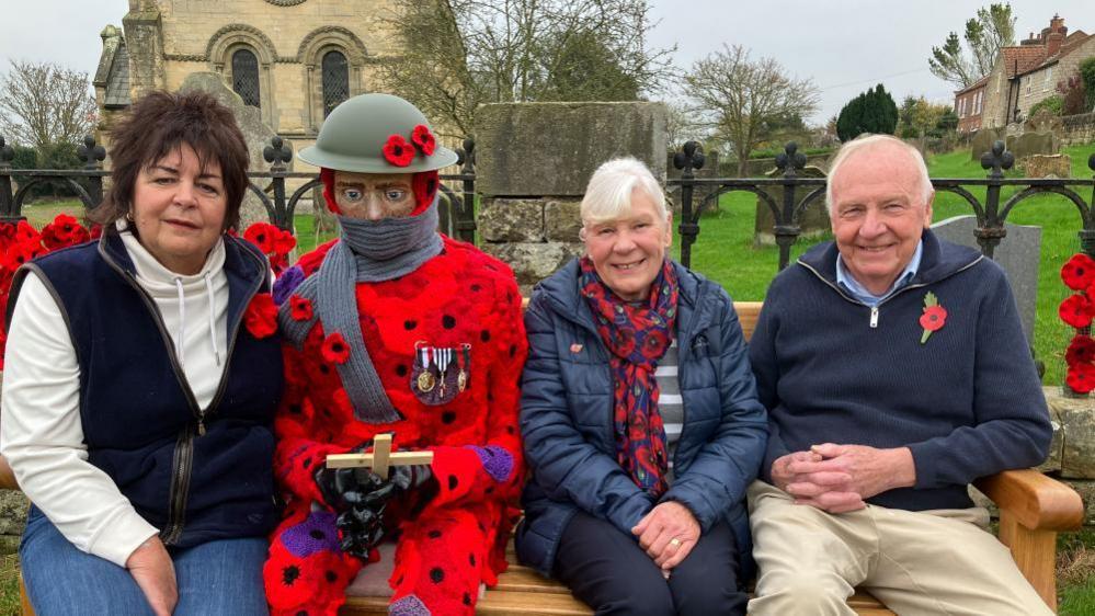 Creators Linda Cook, Susan Hall and Bill Bishop sit next to "Billy Barton" on a bench outside the village church in Barton-le-Street.