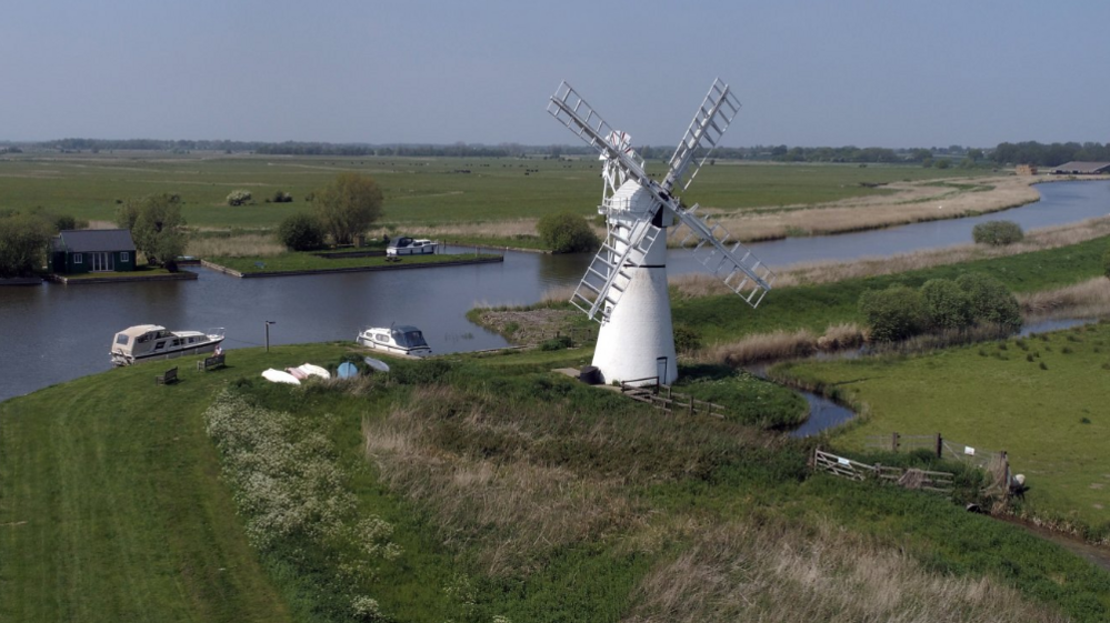 A white windmill on the banks of the River Thurne, with two day boats on the water, surrounded by Broads flatlands and fields.