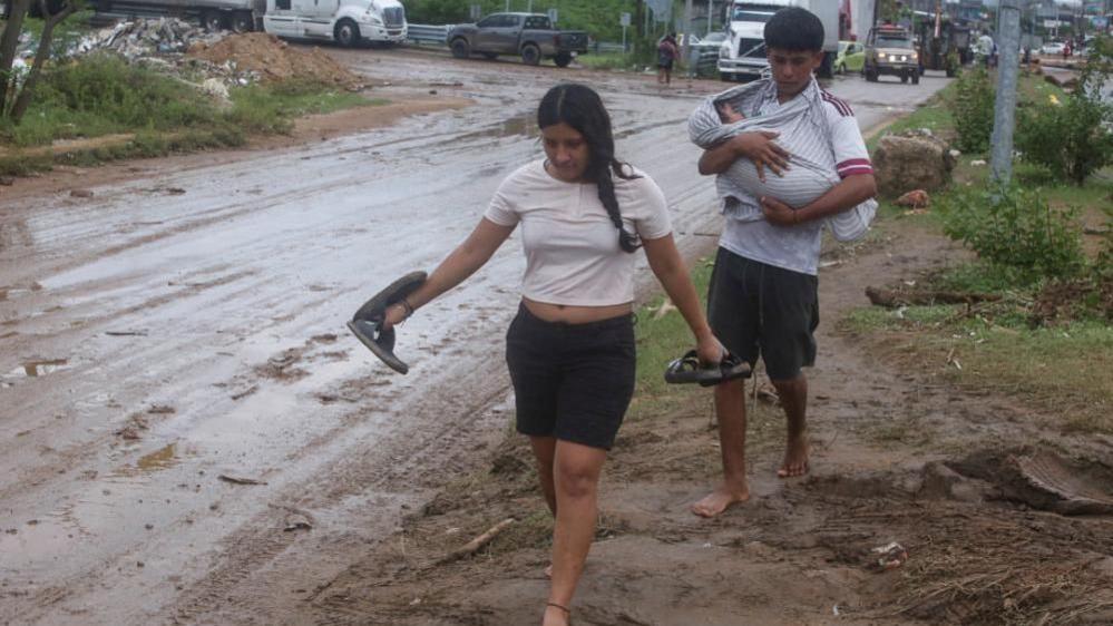 A woman and a man holding a baby walk barefoot on a muddy street in Acapulco, Guerrero, Mexico