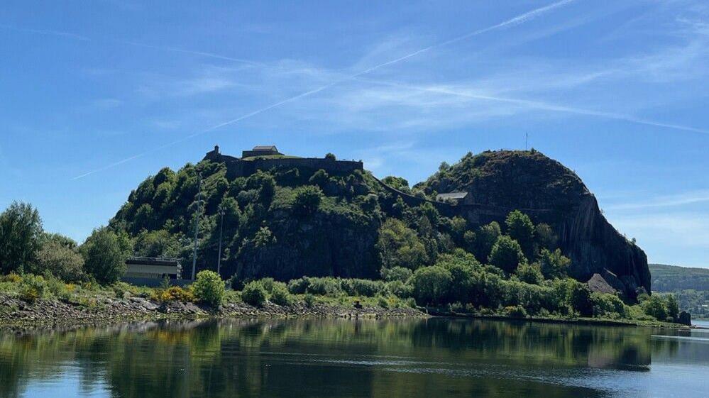 Two steep hills of Dumbarton Rock covered in bushes and trees in the sunshine. The sky is blue and vapour trails