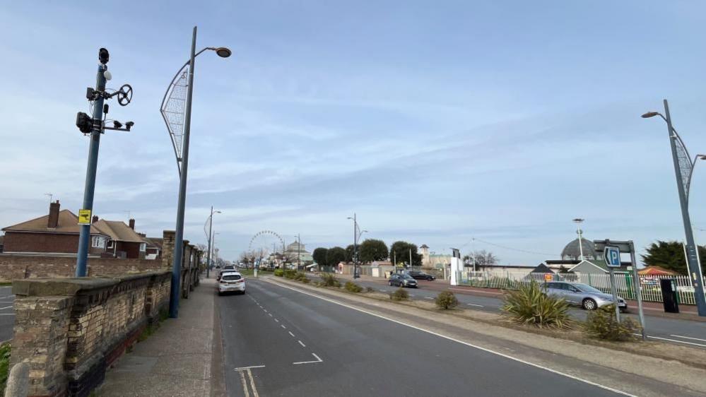 Great Yarmouth's Marine Parade showing a two-lane road with plants separating each lane. Various sea front buildings and plants, with lamp standards each side, plus the camera post with the Accoustic Recognition Camera on the left-most of the picture.