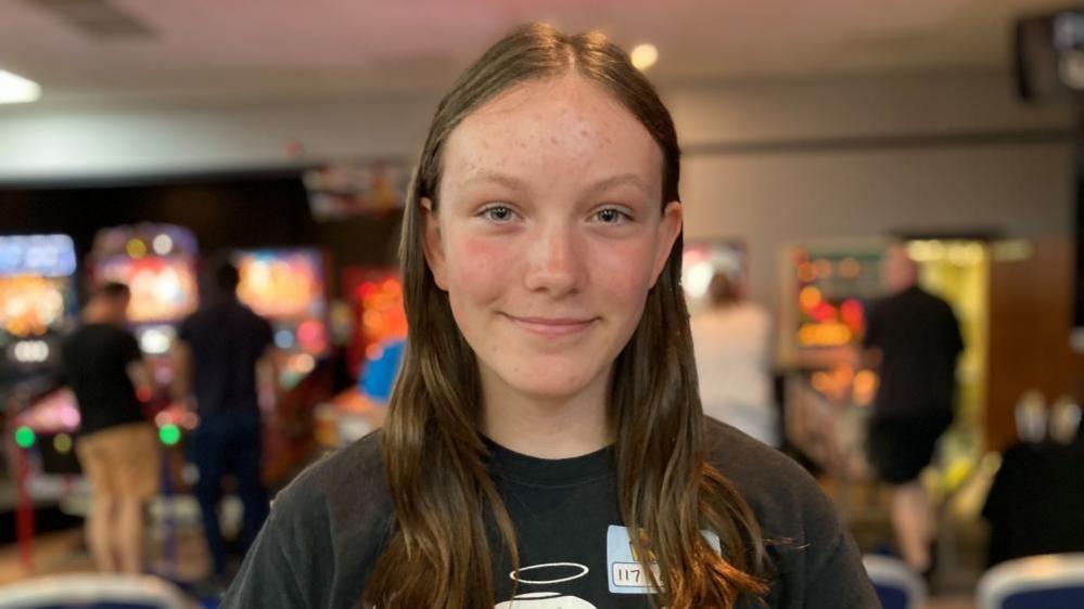 Lucy with long brown hair wearing a black top with pinball machines behind