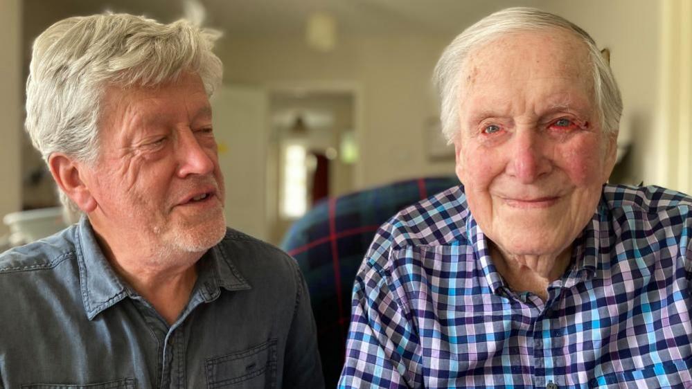 Richard, looking at his father, Leslie. He is wearing a blue plain shirt. His father is wearing a blue checked shirt. They both have short grey hair and are smiling. 