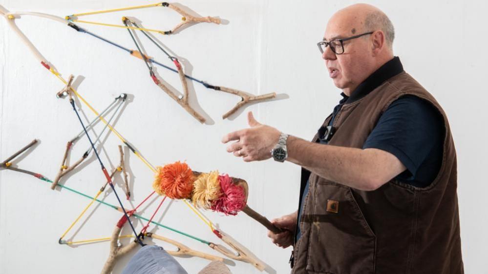 A man with black glasses and wearing a brown gilet holds a handmaid craft. In the background catapults are presented on the wall in odd directions. He holds a hand out and appears to be explaining something. 