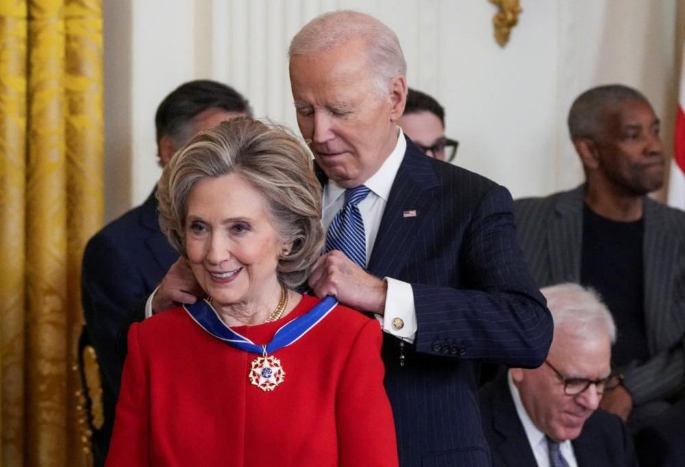 Joe Biden places a medal around Hillary Clinton's next as she wears a red suit