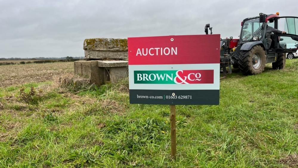 For sale board, which reads "Auction Brown and Co" and gives contact details: brown-co.com 01603 629871, with concrete access to left of the sign, and a fork lift telehandler to the right, set within a field