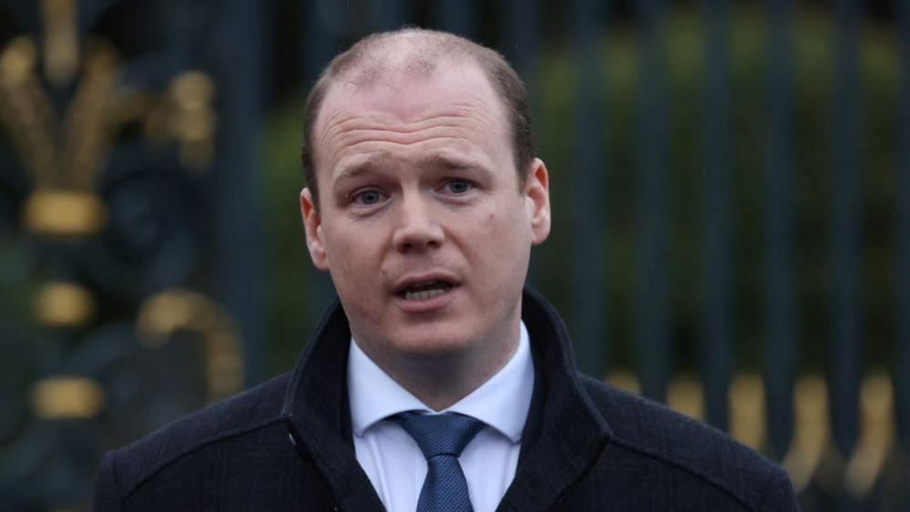 DUP MLA Gordon Lyons dressed in a black coat, white shirt and blue tie in front of a black metal fence. 