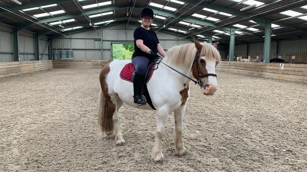 Victoria Cheshire sat on a brown and white horse in an indoor arena looking at the camera.