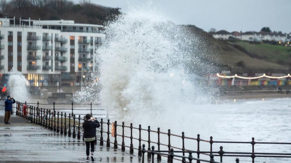 Woman standing on quayside photographing large wave coming towards her