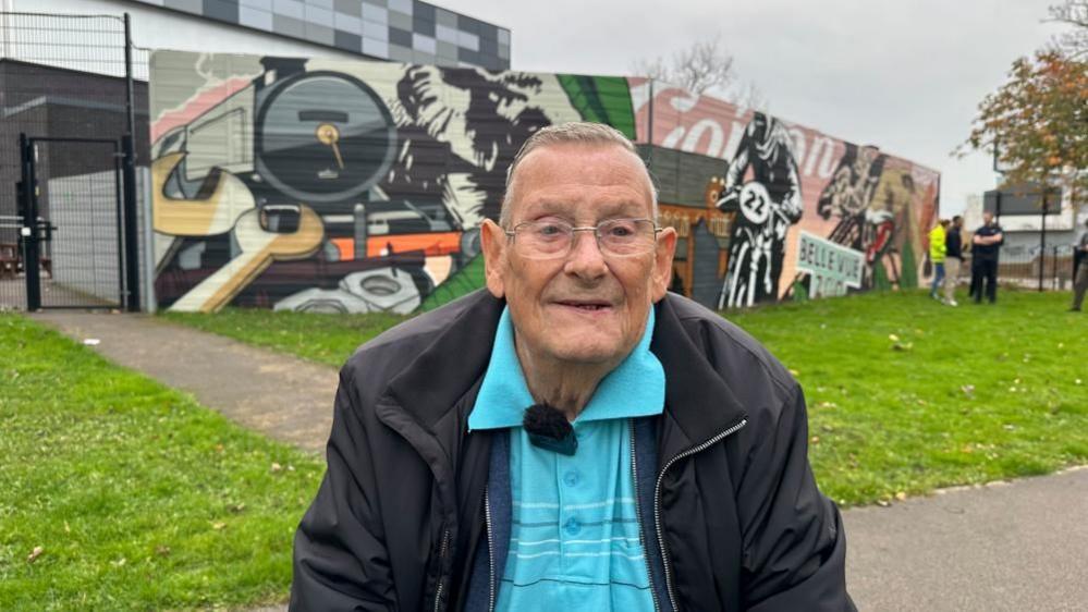 A older man wearing glasses, a blue shirt and a black jacket sits in front of a colourful mural in a park. 