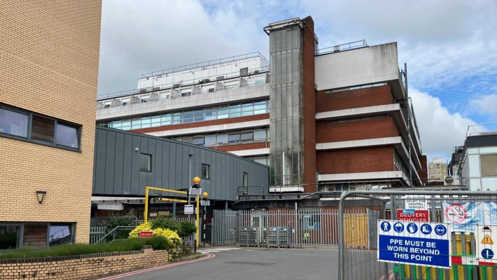 Four-storey concrete hospital building in the background with a first-floor walk way to a modern brick building in the foreground. Metal fencing with a PPE warning is also visible.