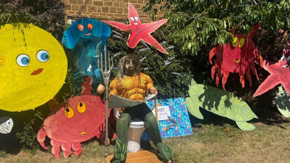 An Aquaman scarecrow sitting on a white toilet holding a silver trident. Around him are a variety of cardboard sea creatures including crabs, puffer fish and a turtle. 