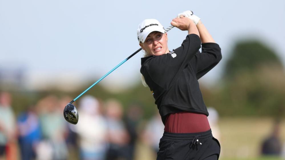 Charley Hull at the end of her backswing. She is holding the gold club behind hand and has a grimace on her face. She is wearing black trousers, a red top with a black jumper over it, as well as a white cap.