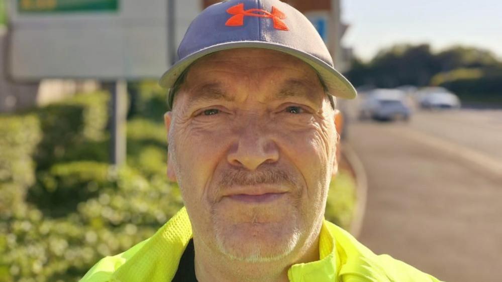 Darren Baker wearing a blue cap and yellow jacket in front of a road sign