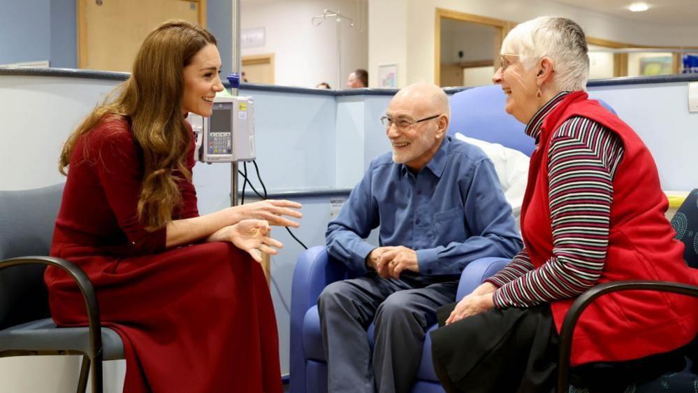 The Princess of Wales speaks to an elderly man and woman who are smiling as they talk with the princess inside the Royal Marsden Hospital