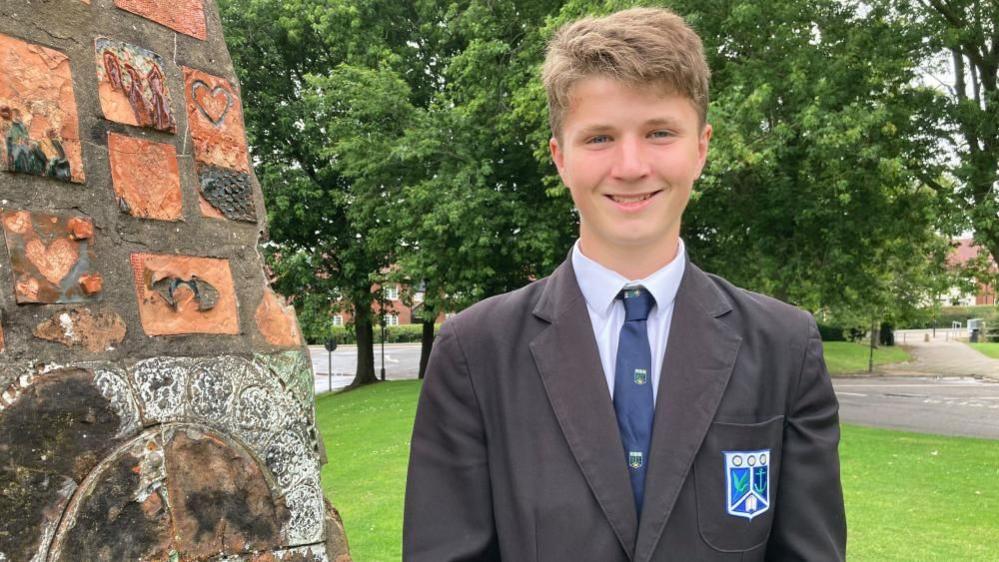 Harry smiles at the camera and is wearing a dark school uniform, including a tie and blazer. He is standing in school grounds by a brick structure