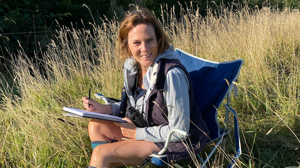Hannah Dale is sitting in a camping chair in a grassy field, she is holding a notebook and smiling at the cameras. Hanging from her neck is a pair on binoculars, she is also wearing a grey jacket.