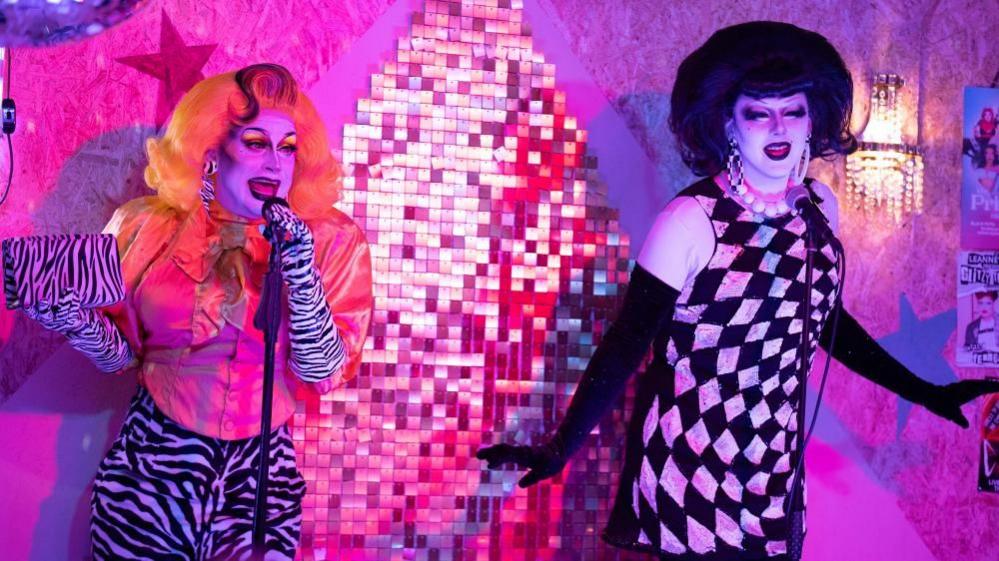 Anna Phylactic stands next to fellow drag queen Liquorice Black. Anna wears leopard print gloves and trousers and is wearing a fluro orange wig. Liquorice Black wears a black wig and a black-and-white dress.