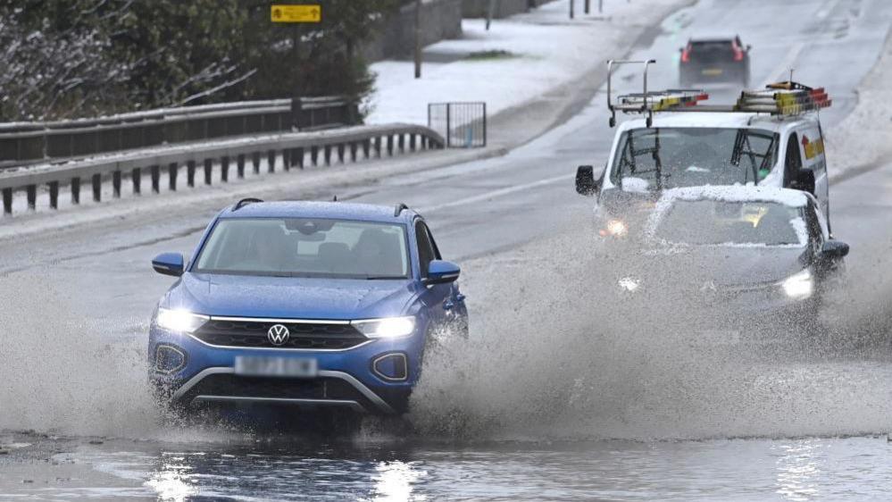 A blue Volkswagen car with its headlights on drive through a shallow flood causing water to splash on both sides. Behind it is a grey car with snow on it and a van. The road in front of the vehicle is flooded while the road behind it are still covered in snow