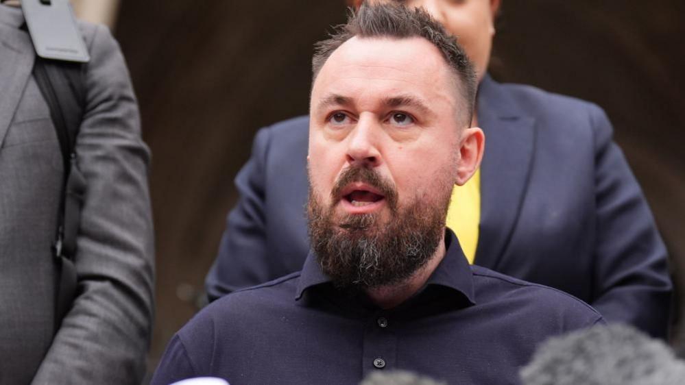 Martin Hibbert sits speaking. A microphone with a fluffy cover is in the foreground. People in suits stand behind him.