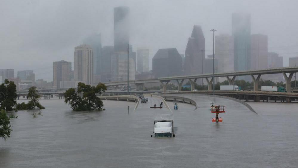 Houston skyline in the background, covered in cloud. In the foreground is a highway running into a large flooded area. A lorry, facing the camera, is partly submerged. There are a few trees on the left-hand side.