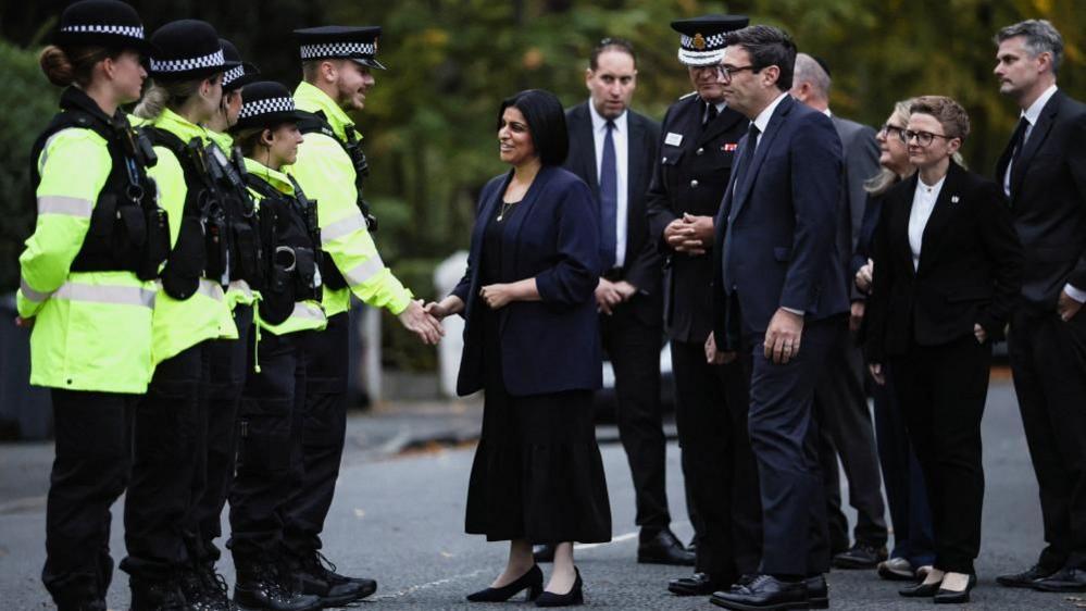 Shabana Mahmood stands shaking hands on a street with a police officer who is standing next to a line of four other police officers in high-vis jackets. Standing behind her is Greater Manchester Mayor Andy Burnham, Manchester Council leader Bev Craig and Greater Manchester Police Commissioner Stephen Watson.
