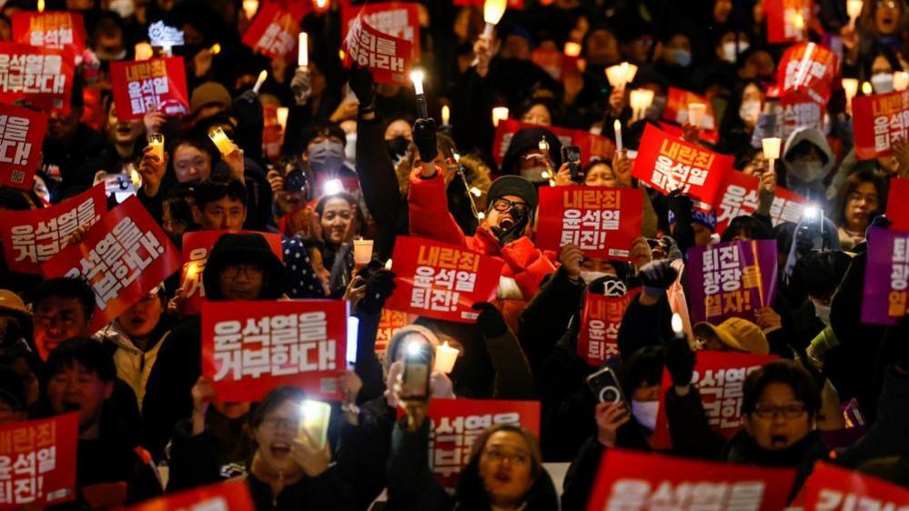 A crowd of protesters fills the frame, holding electric candles and red placards. One man in focus in the centre holds a gloved hand to his mouth as he yells