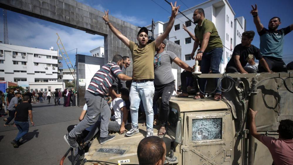 Palestinian stand on a captured Israeli jeep on a street in Gaza (7 October 2023)