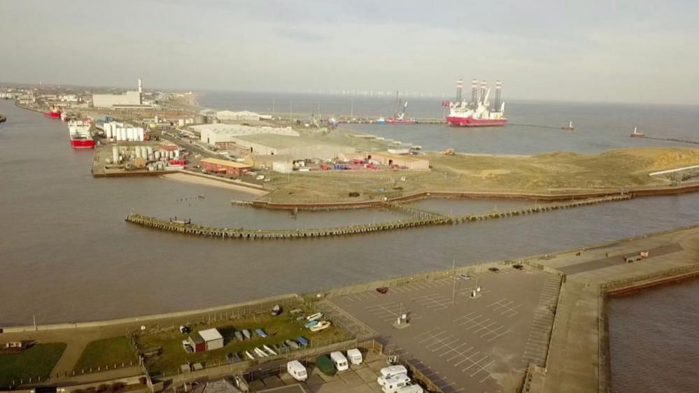 A drone image showing Gorleston pier in the foreground, with the Great Yarmouth river port running out to sea in the centre, the harbour bearing up to the left of the image, and in the distance the outer harbour itself. The undeveloped land is in the centre right of the image with water either side. Various buildings and some ships are visible in the picture.