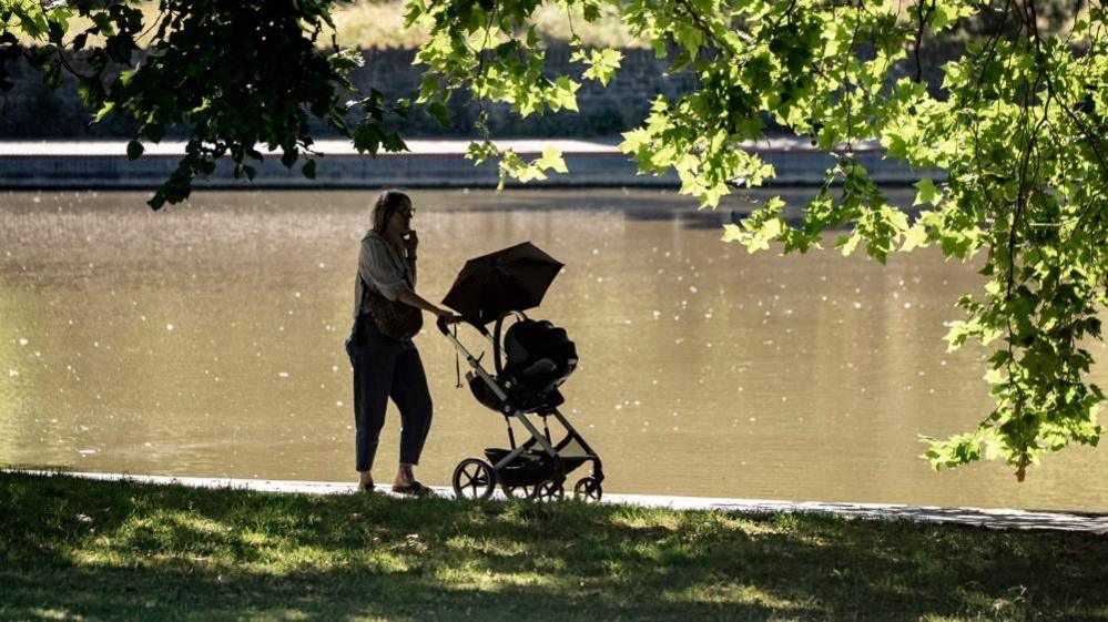 A woman pushing a buggy along a path beside a pond in a park. She and the buggy are both silhouetted by the sun. It's a bright day, with the sun shining off the water and on the leaves of trees.