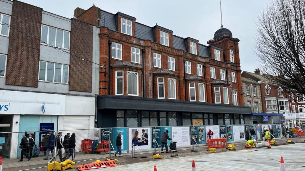 The former Palmers Department Store, with its refurbished windows and facade, and in the near-ground, the new paving of the market place. Hoardings surround the building site and people are walking close by. There are cones and barriers visible.