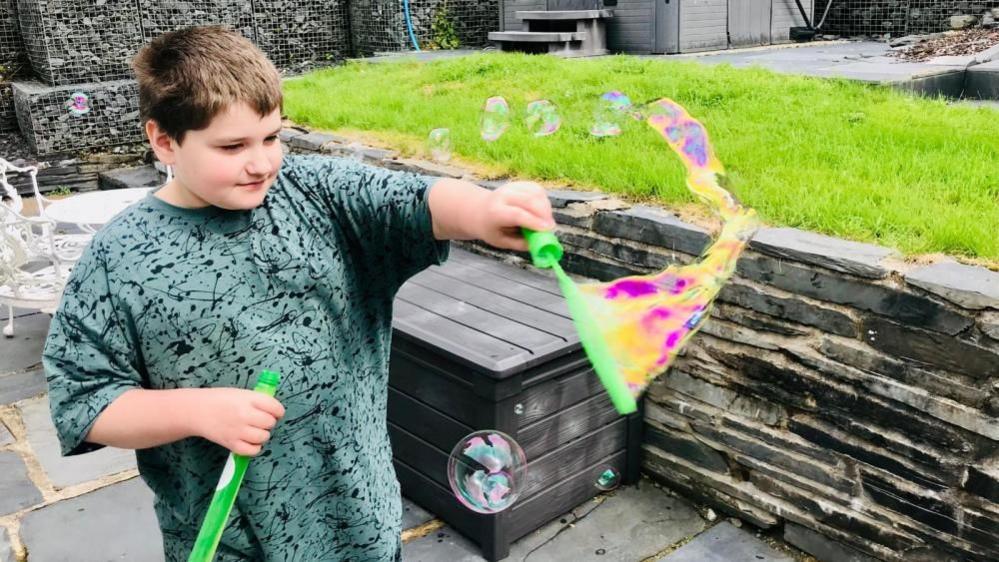 Sam with short brow hair wearing a green T-shirt and blowing bubbles