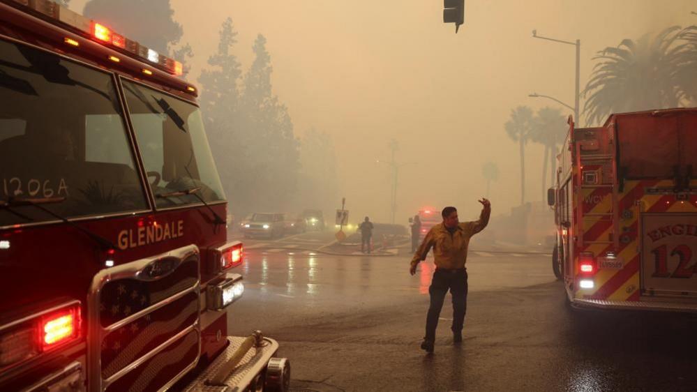 A fire official gestures to oncoming traffic in the centre of a junction. There are fire trucks on either side of him and car headlights shine through thick smoke from the wildfires.