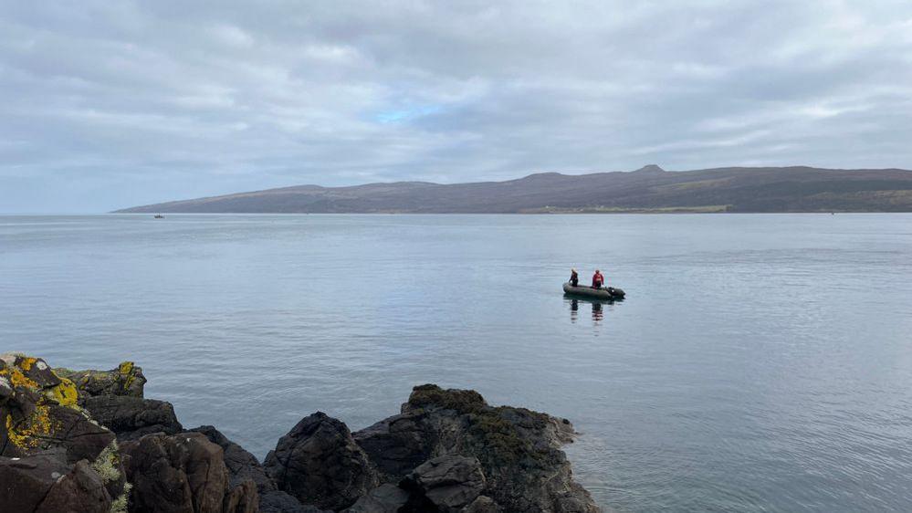 Two men in an inflatable boat on clear, quiet waters. Land can be seen in the distance and the weather is cloudy but calm.