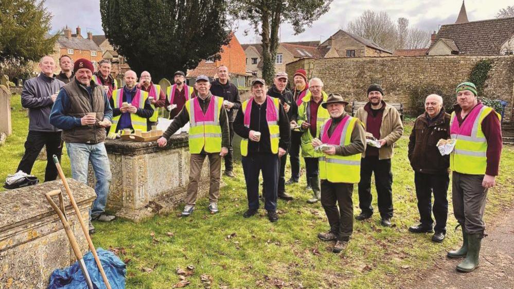 Several people are stood in hi-vis jackets holding food and drink in a churchyard.