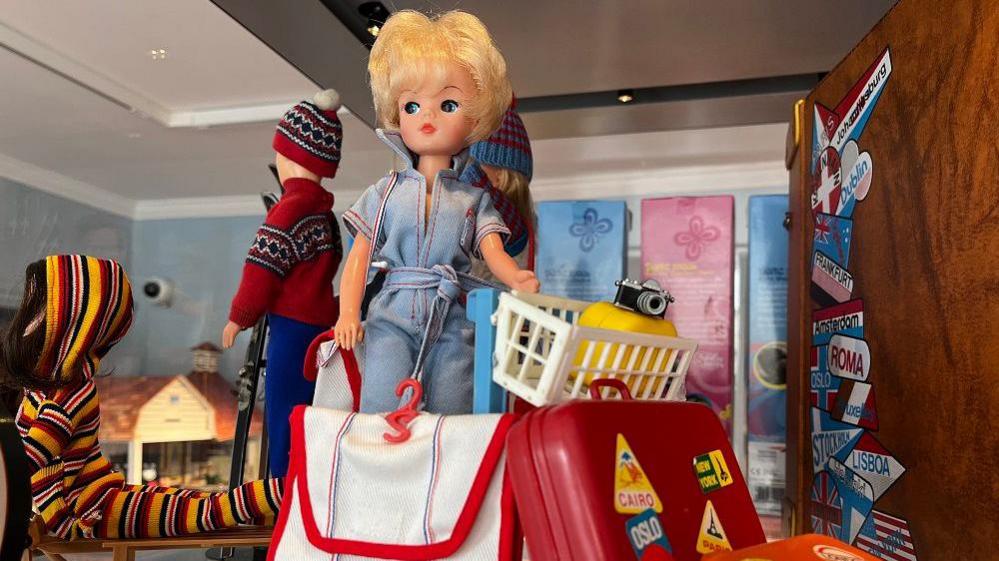 A Sindy doll wearing a jumpsuit and with a luggage trolley of cases