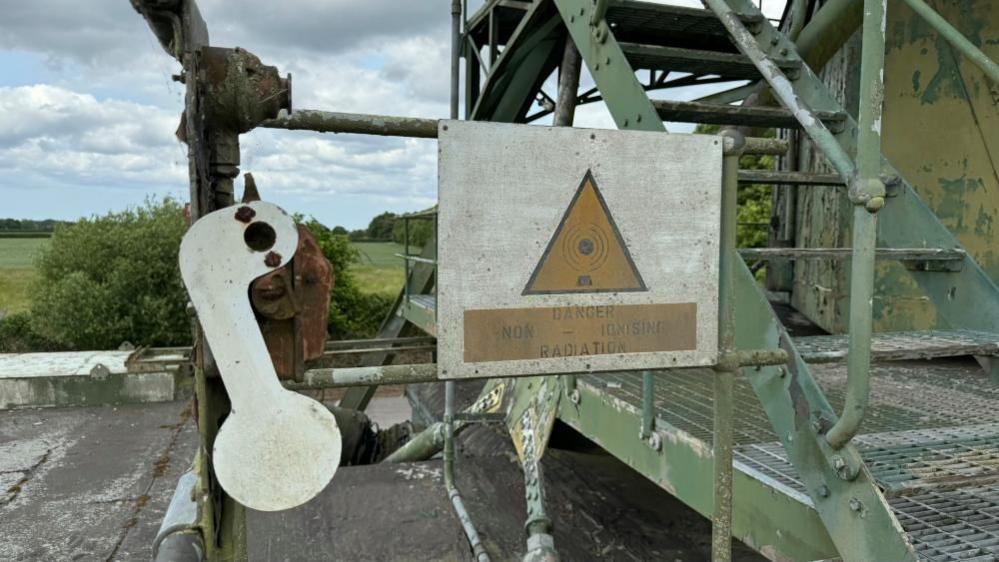 A white shape with coloured lens aperture, and a warning sign that states: "Danger; non-ionising radiation" and a yellow warning triangle mounted onto green-painted hand rails on a staircase up the T84 radar.