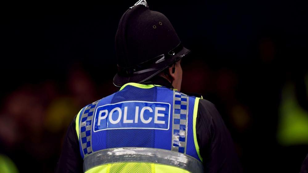 A police officer wears a high vis vest which says "police" on it as well as a helmet and faces away from the camera. 